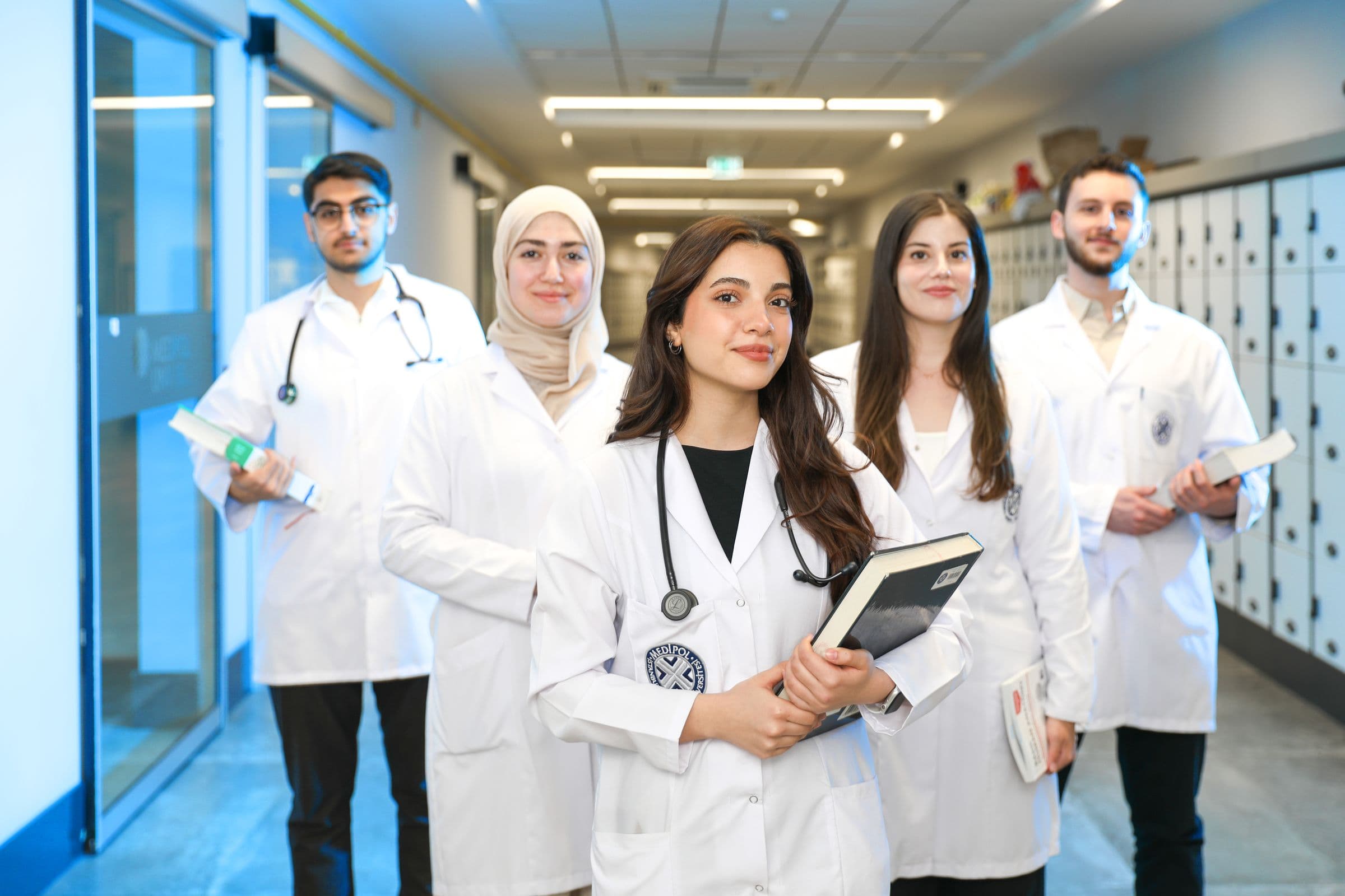 Five medical students in white coats with stethoscopes and textbooks, standing in a university corridor