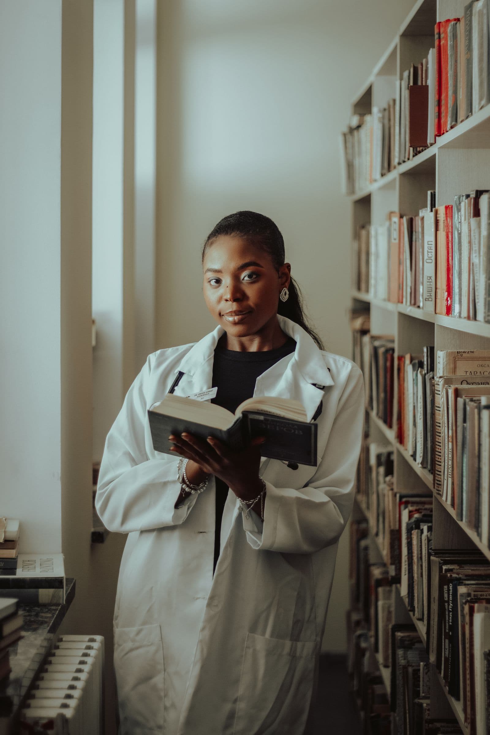 A medical student in a white coat reading a book beside library shelves